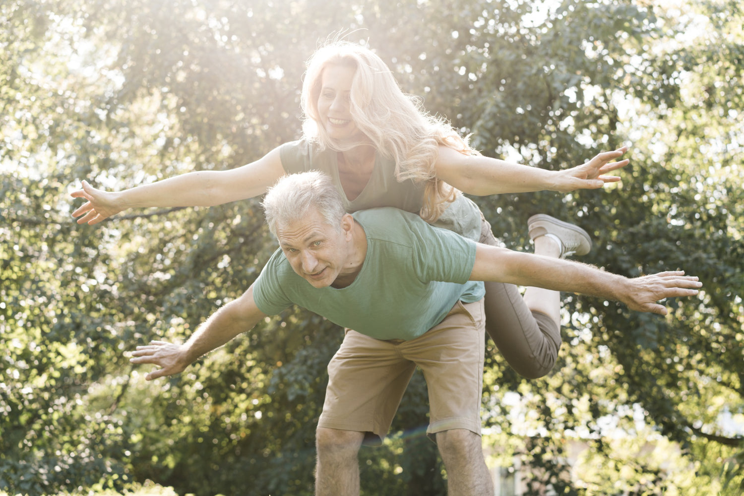 A happy, energetic older couple playing outdoors in a park. The man is giving the woman a piggyback ride as they both spread their arms like airplane wings, smiling joyfully. Sunlight filters through the trees, creating a warm, healthy atmosphere.