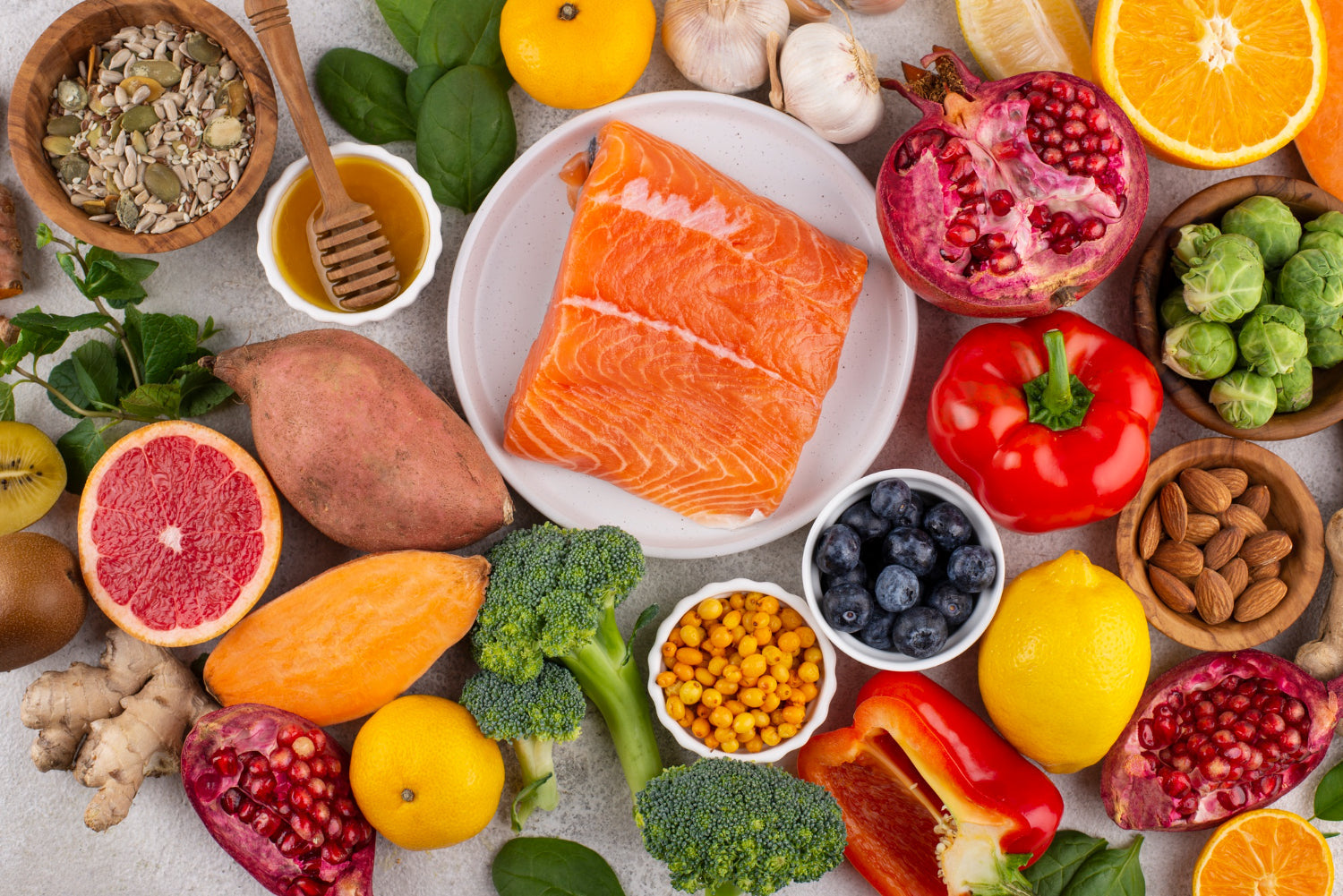 A variety of gut-healthy foods arranged around a plate of fresh salmon, including pomegranate, blueberries, sweet potatoes, broccoli, almonds, red bell pepper, garlic, spinach, citrus fruits, seeds, honey, and ginger.